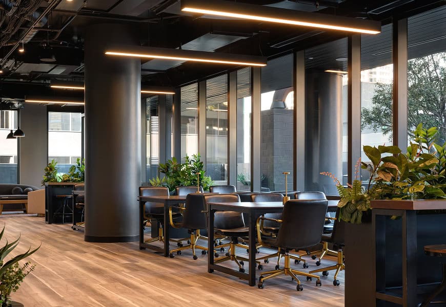 Coworking space with tables, chairs, plants, glass walls, and exposed ceiling.