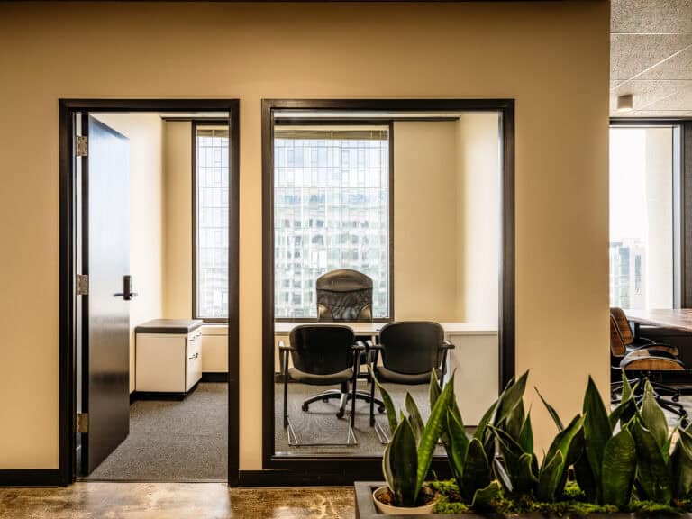 Glass-walled office with desks, chairs, and plants near interior windows.