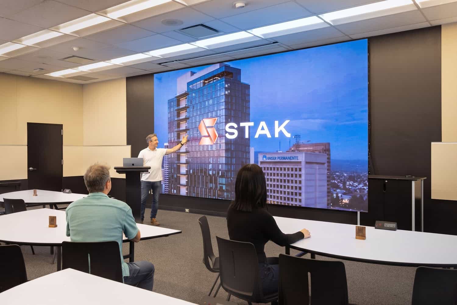 Presentation room with attendees at tables facing a large STAK screen.